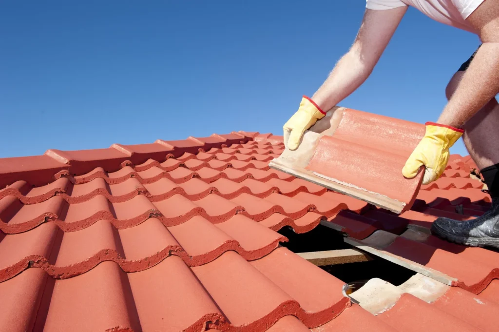 Person wearing gloves replacing a red roof tile on a tiled roof under a clear blue sky, with a visible hole where tiles are missing.