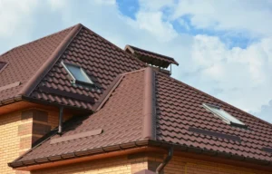 A close-up view of a house roof with brown metal roofing tiles, two skylights, and brick exterior walls against a partly cloudy sky.