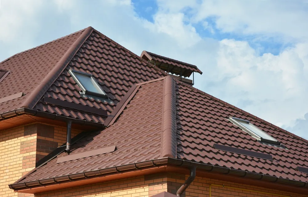 A close-up view of a house roof with brown metal roofing tiles, two skylights, and brick exterior walls against a partly cloudy sky.