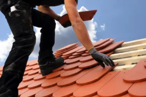 A person installs terracotta roof tiles onto wooden beams, wearing gloves and using a tool under a partly cloudy sky.