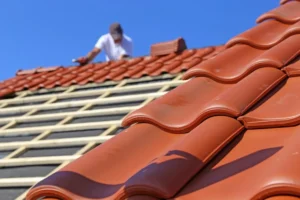 A person installs red clay roof tiles on a wooden framework under a clear blue sky.