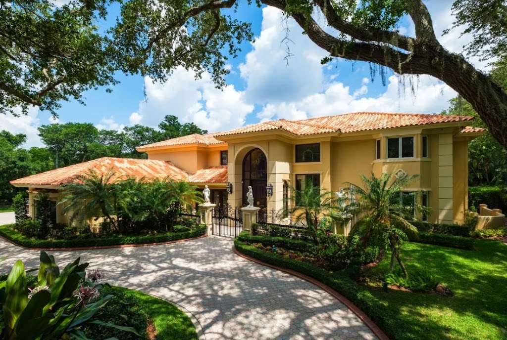 Large two-story yellow house with a red-tiled roof, arched entryway, palm trees, and a curved driveway, surrounded by lush green landscaping—perfect for enjoying warm weather under a partly cloudy sky.
