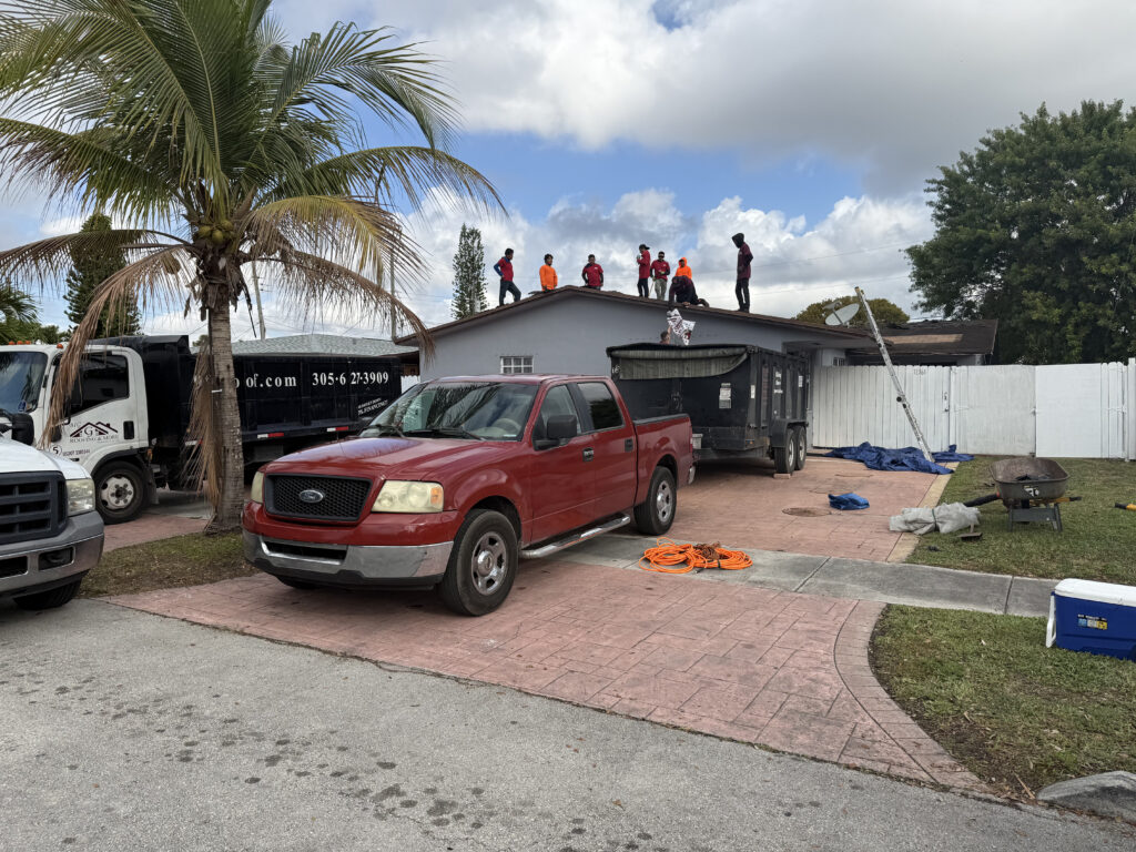 A group of metal roof installers stands on the roof of a single-story house, with trucks and equipment parked in the driveway under a partly cloudy sky.