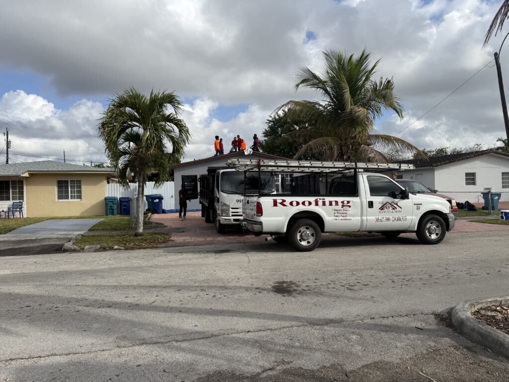 A roofing truck is parked in front of a house while several workers install a new roof. Two palm trees are in the yard, and the cloudy sky hints at a Florida storm—reminding you to protect your home with regular roof inspections. location Miami.