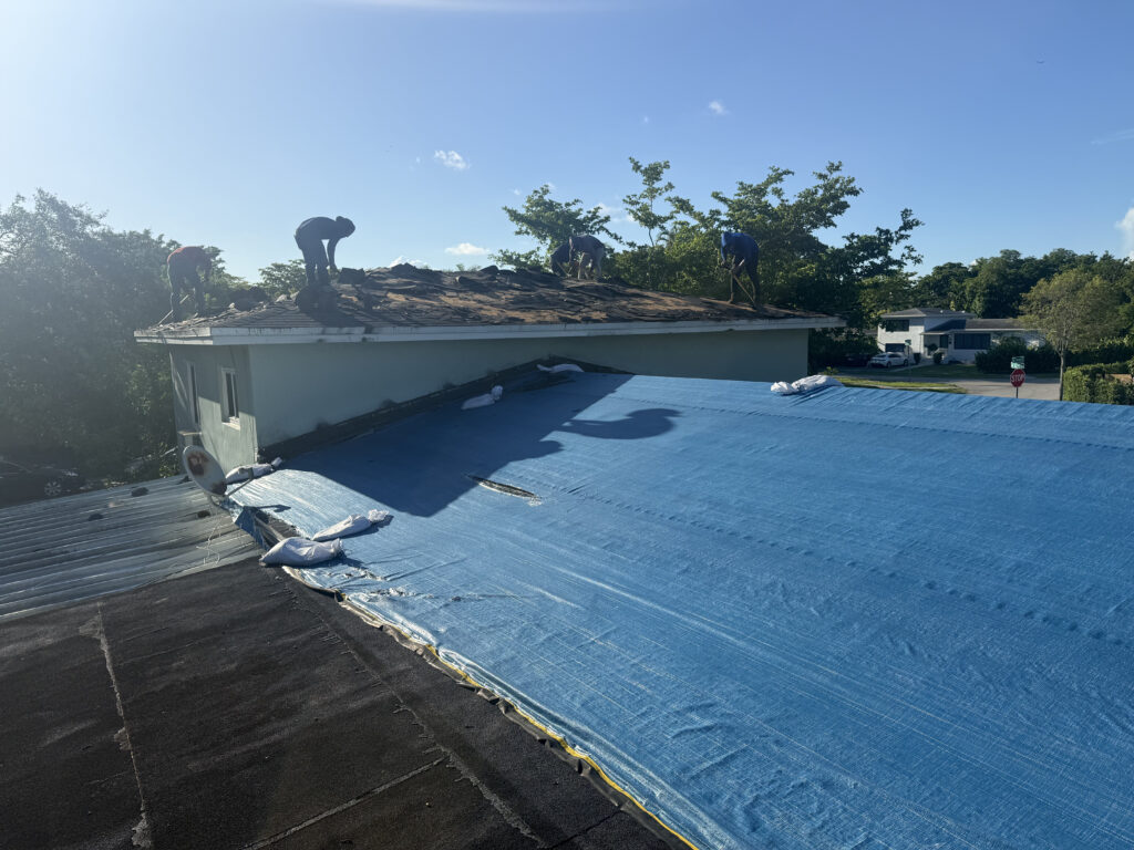 Workers remove old shingles from a house roof, while a large blue tarp covers the adjacent roof section on a sunny South Florida day—reminding homeowners of the importance of regular roof inspections.