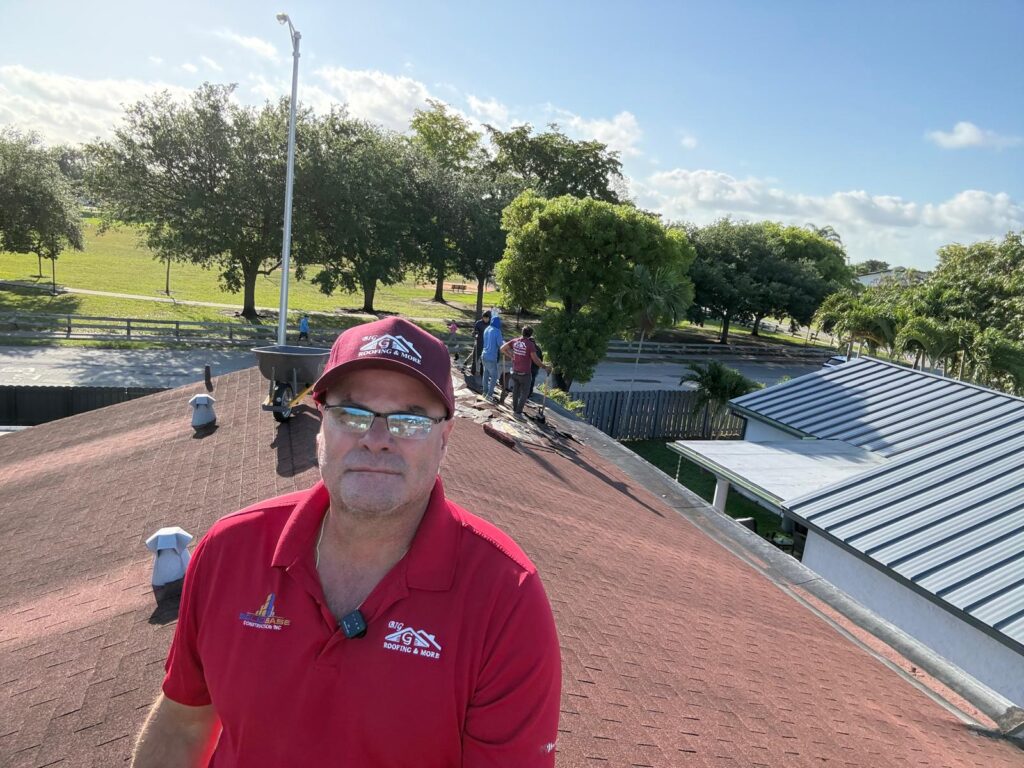 Man wearing glasses and a red hat stands on a red shingle roof, showcasing effective roof repair; several other people work in the background, with trees and a road visible beyond the building.